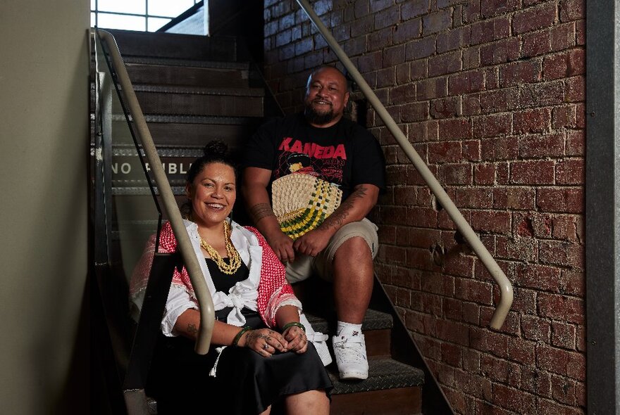 Man and woman seated on steps in a stairwell with brick wall and railings.