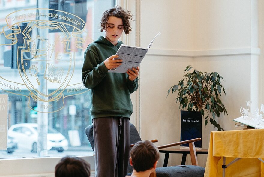 A child standing up in front of class reading with a large window behind them. 