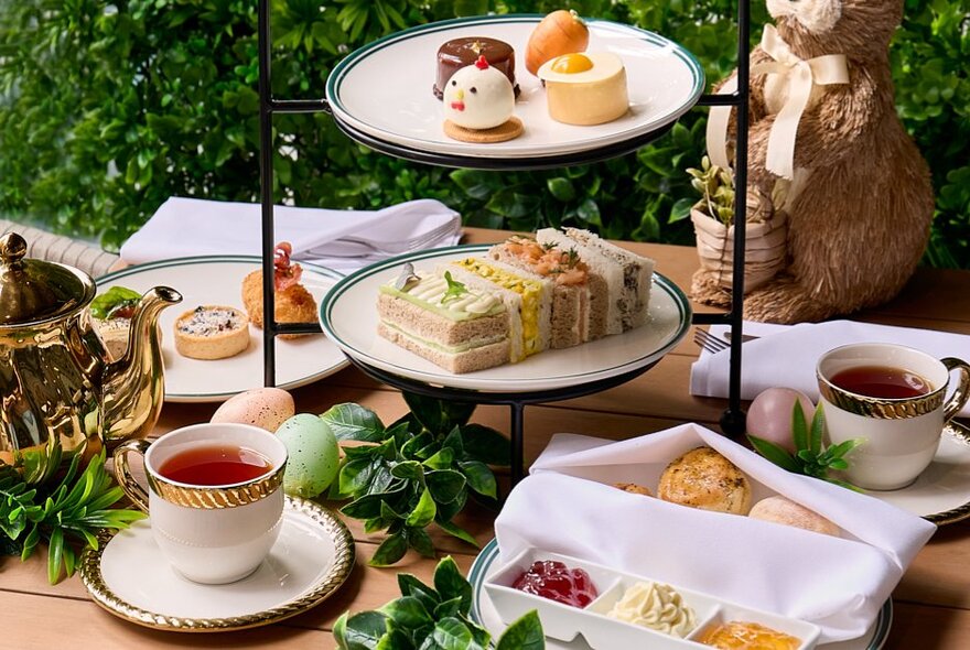An Easter themed high tea served on tiered stands, with small portions of sweet and savoury food on display, a pot of tea, cups and saucers, and an small bunny sculpture in the background. 
