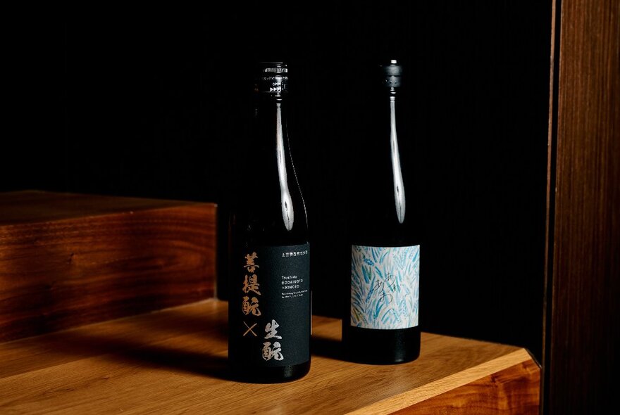 Two black bottles of Japanese sake resting on a wooden bench; a black wall behind them.
