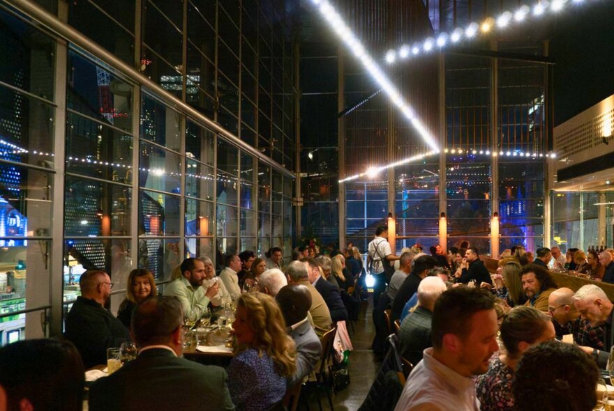 Airy dining space at Taxi Kitchen, at night, with the dining space filled with patrons seated at tables eating and drinking.