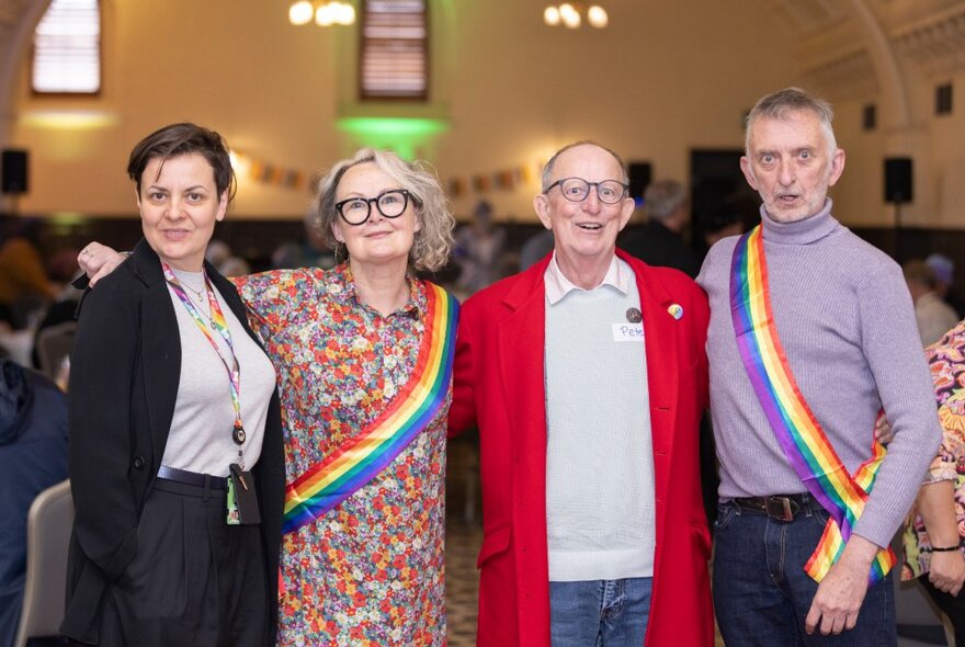 Four older people lined up in a row and smiling, two of them wearing rainbow sashes across their chests.