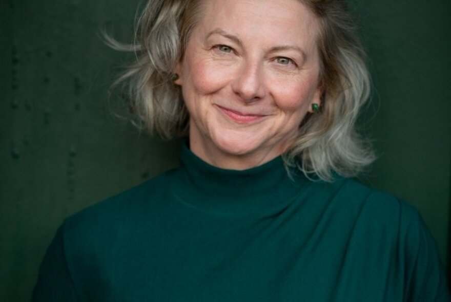 Author Toni Jordan, smiling directly at the viewer, with shoulder length ash blonde hair, wearing a dark green jumper, against a lighter green background.