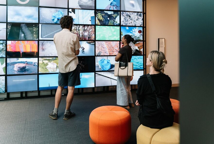 People standing in front of a wall of small screens, each displaying a different image, a woman sitting on a large cushion seat. 