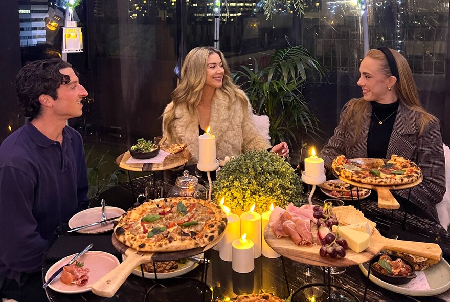 Three patrons enjoying a meal with pizza, charcuterie boards and candles on the table in front of them.