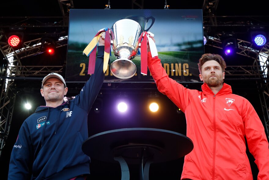 Two AFL footballers holding up the Grand Final premiership cup on a stage at Yarra Park.