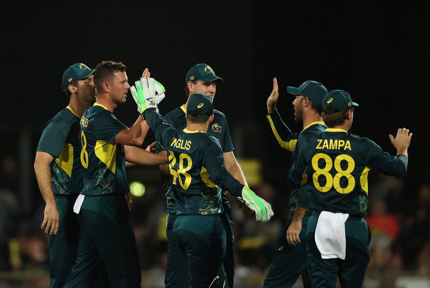 Australian men's cricket team wearing dark green and yellow uniforms, celebrating with high fives and claps, in a huddle on the sports field.