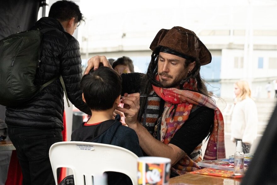 A child getting his face painted by a man dressed as Captain Jack Sparrow.