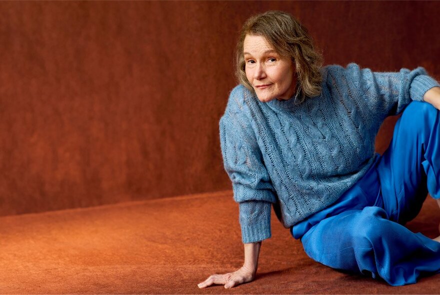 Actor seated on an orange carpeted surface, wearing a blue jumper and trousers.