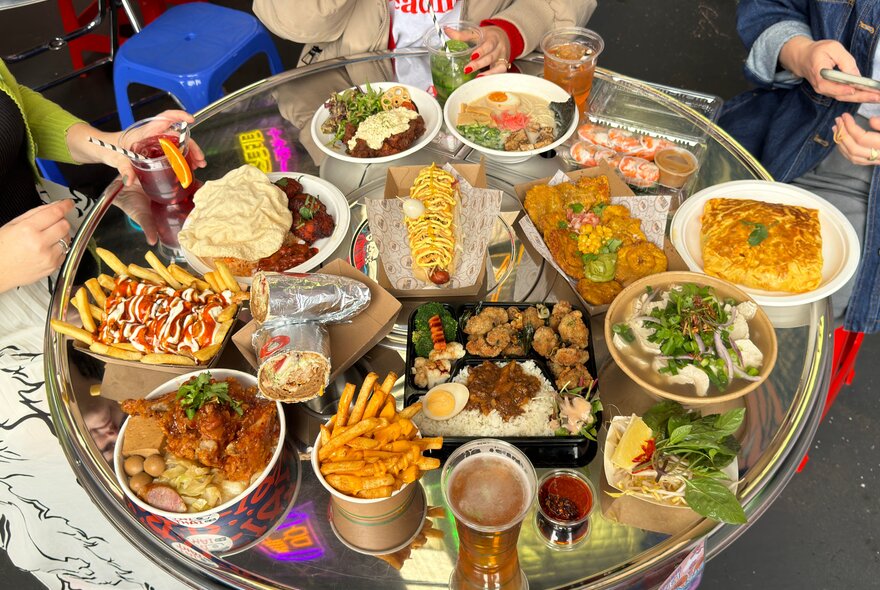 An aerial view of a metal table with many dishes and drinks laid out, including chips, noodles, a kebab, fried chicken, a hot dog, a bento box, ramen, rice paper rolls, pho and more.