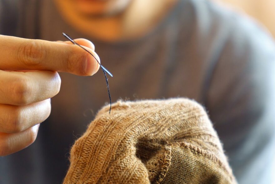 Hands holding a needle and thread over an item of woollen clothing.