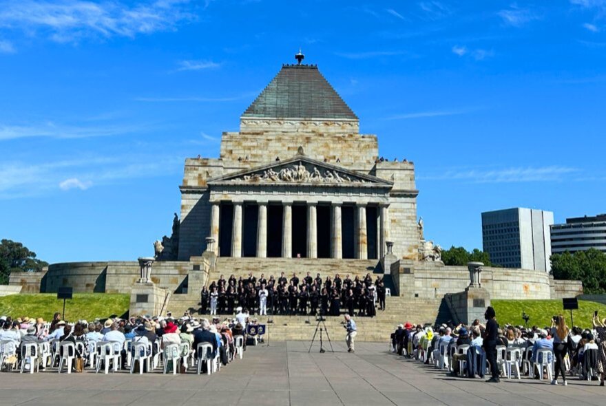 People seated in chairs on the forecourt in front of the Shrine of Remembrance, listening to chorus of singers who are standing on the steps of the Shrine.