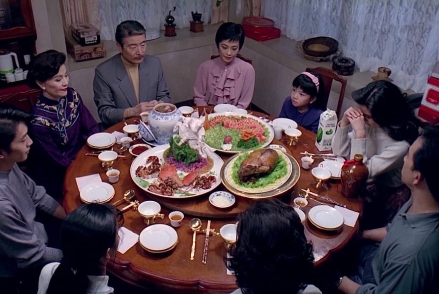 Looking down at a family seated at a round dining table with three large platters of food and white crockery; a still from the film, Eat Drink Man Woman.