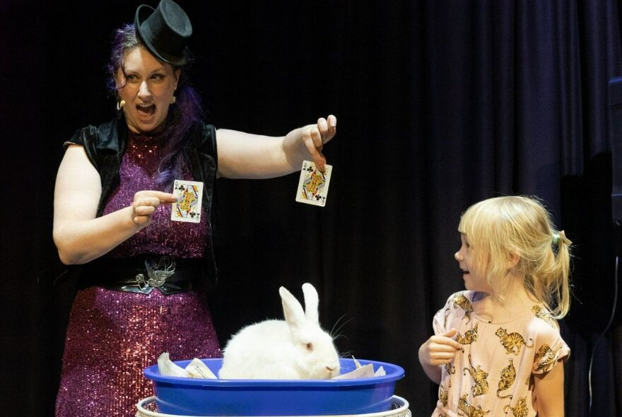 Magician Lee Cohen holding two playing cards in each hand, standing in front of a white rabbit on a stage, with a young child standing on the side of the stage and watching.