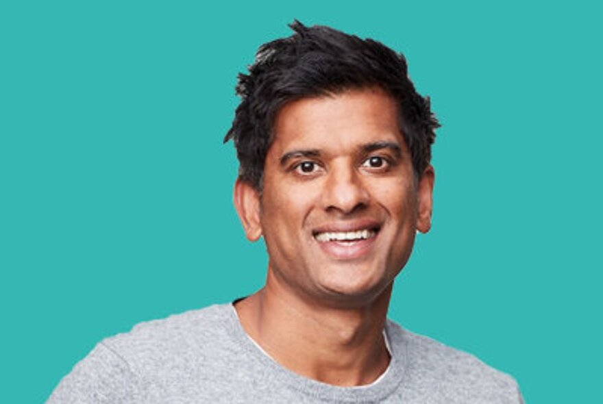 Smiling Indian man wearing a grey jumper against a turquoise background.