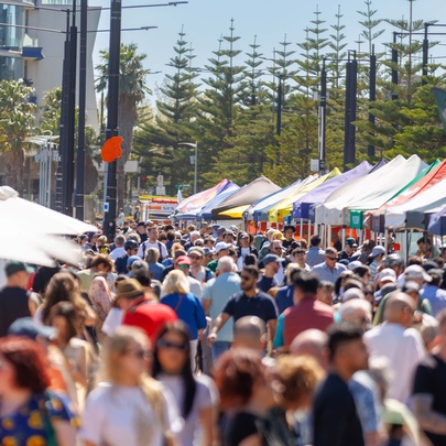 A crowded outdoor market on a sunny day, with numerous people walking down a path lined by market stalls and umbrellas under a blue sky.