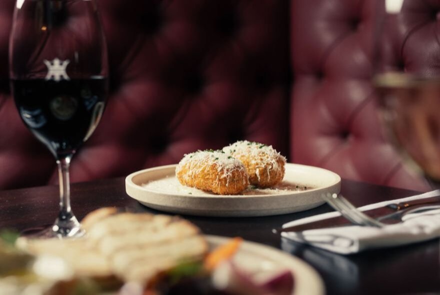 A plate of food and a glass of red wine served in a restaurant setting, burgundy banquettes in the background.