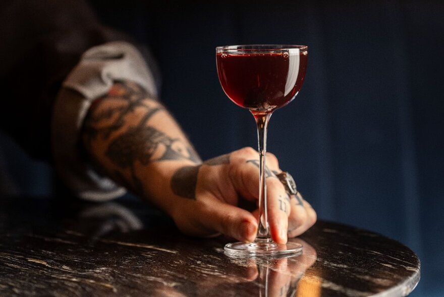 A tattooed hand touching the stem of a small glass filled to the brim with deep red liquor. 