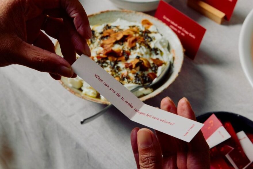 Over a meal, a person unfurls a printed message with a question about being a refugee.