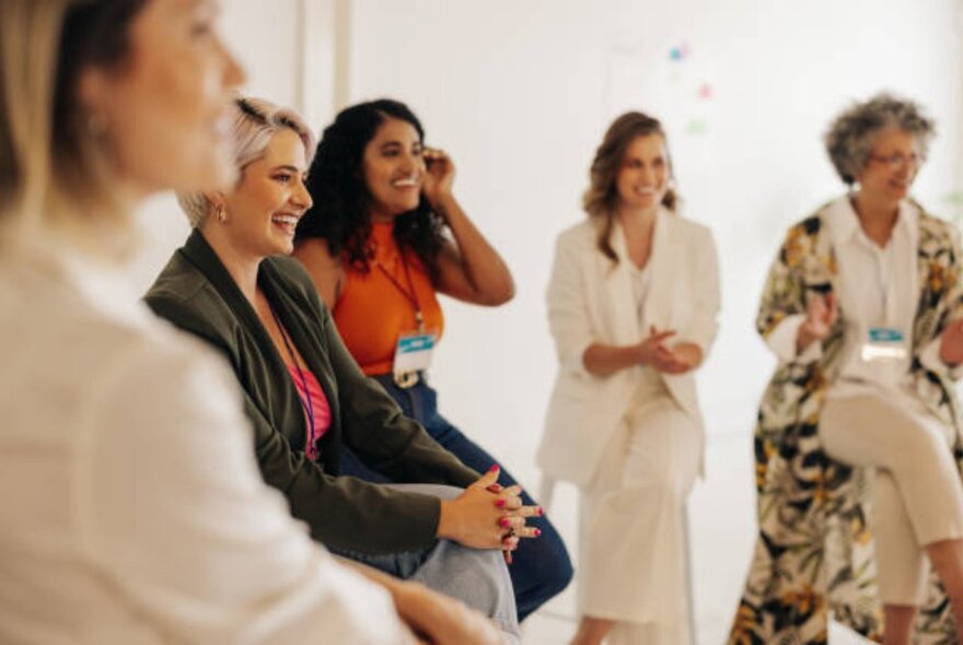 A diverse group of well-dressed women smiling in a casual setting and looking at something out of shot.