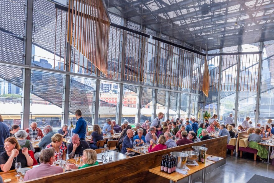 The interior of Taxi Kitchen at Transport Hotel, with tables of patrons eating and drinking at long tables, surrounded by high floor to ceiling windows.