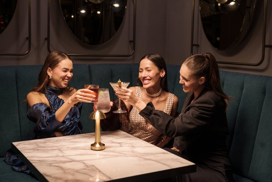 Three people clinking glassess over a marble table in a dimly lit room.