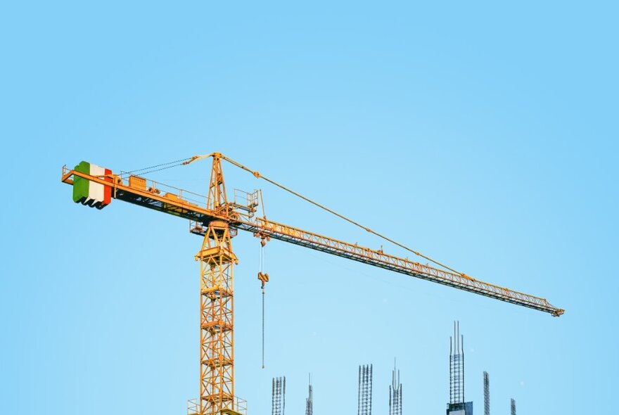 A large yellow crane with a counterweight painted with the colours of the Italian flag, against a bright blue sky with buildings in the background.