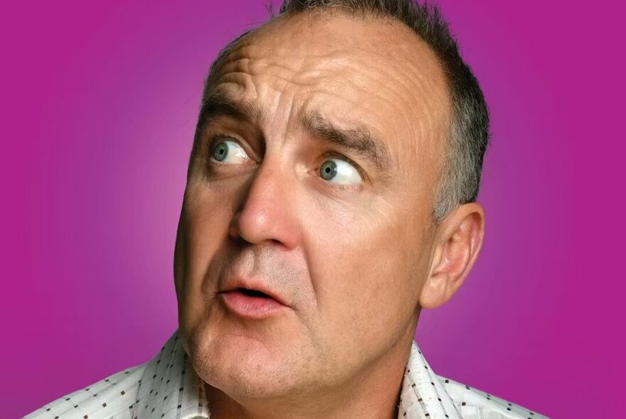 A close-up of comedian Jimeoin with a surprised expression, eyes wide and mouth slightly open, looking to the left against a magenta background.
