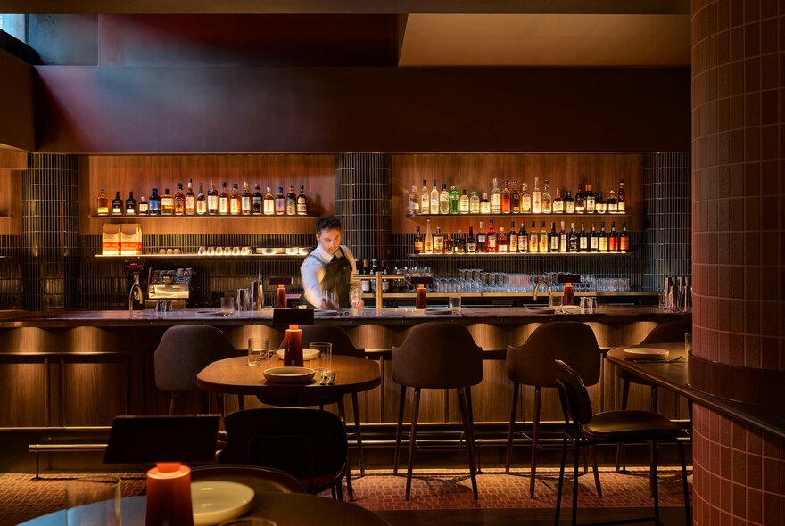 Interior of a bar, with a bartender behind a timber counter, a wall of bottled drinks behind him, high bar stools and some tables in the foreground.
