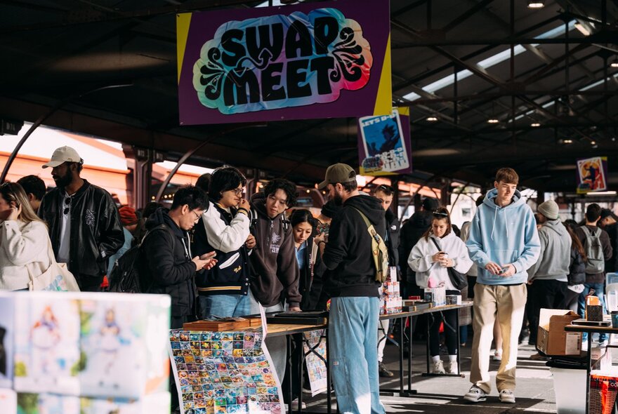 Stalls at the Queen Vic Market selling collectables, with people walking around and browsing, with a large sign hanging from the shed ceiling that reads SWAP MEET.