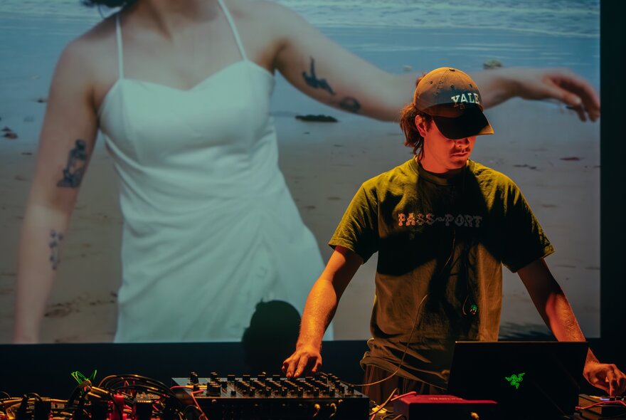 A DJ working the decks in front of a screen showing a woman in a strappy white dress on the beach.