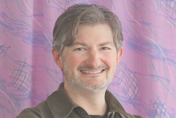 Smiling middle-aged man, with short greying brown hair, posed against a pink background.