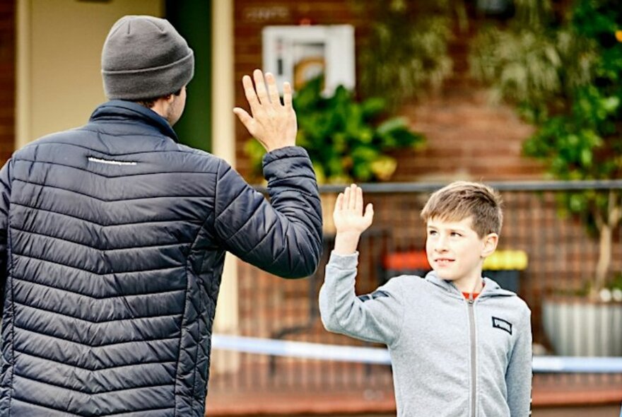 An adult and young boy high-fiving in front of a tennis court net.