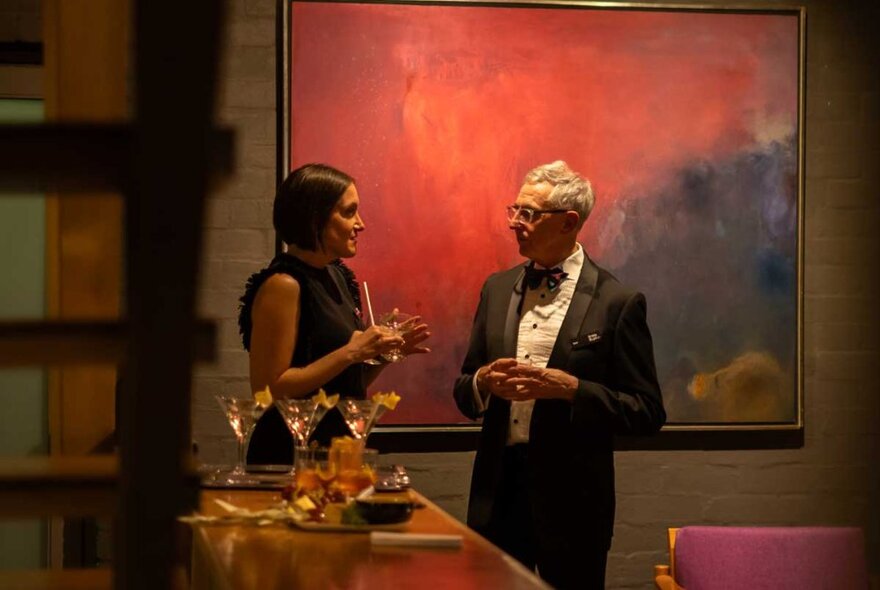 A well-dressed couple talk over cocktails in a room with modern artwork on the wall. 