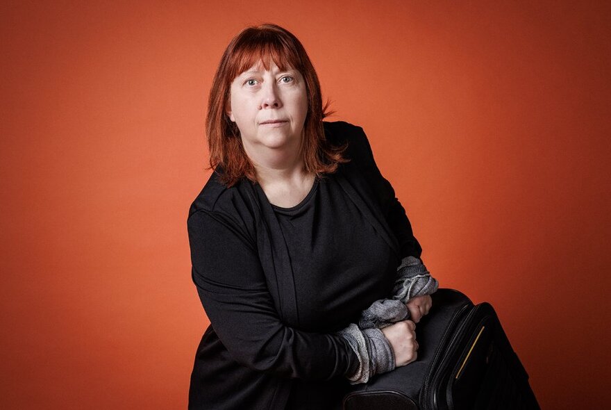 A woman looking concerned at the camera, wearing a black outfit and holding a black suitcase in front of a red background.