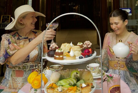 Two ladies in period costume sharing a high tea with cakes and a teapot.