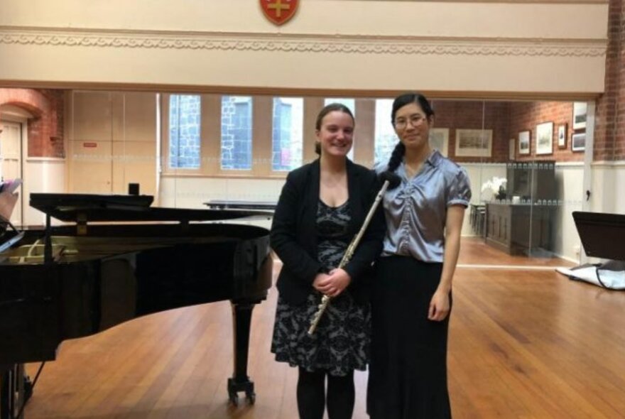 Flute and piano duo, Kelsy De Prada and Rosa Hwang, posing in a music studio in front of a grand piano.
