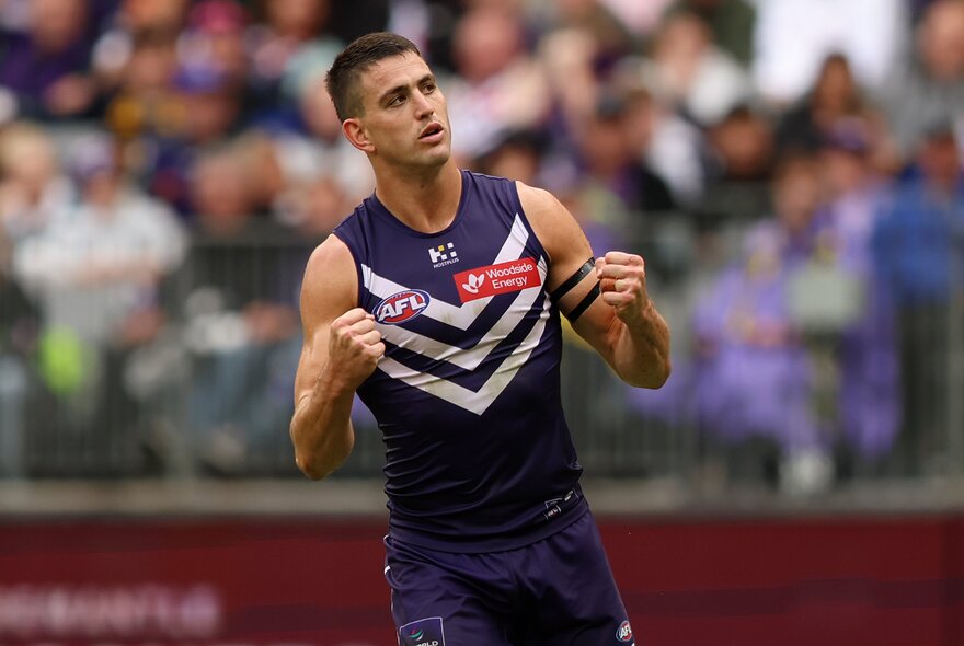 A Fremantle Dockers AFL player celebrating on the field with his fists clenched and up near his chest, a blurred stadium crowd behind him.