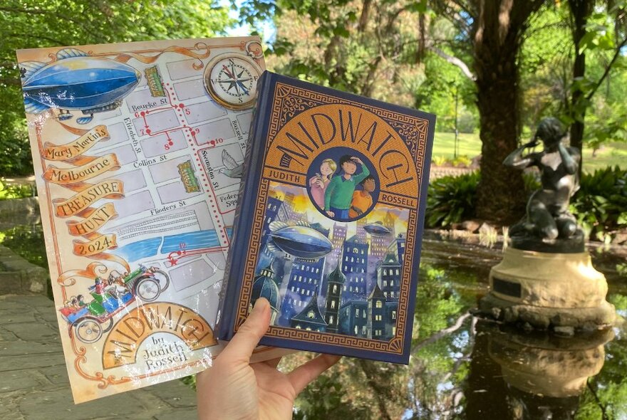 A hand holding up a map and a book in a garden setting near a lake.