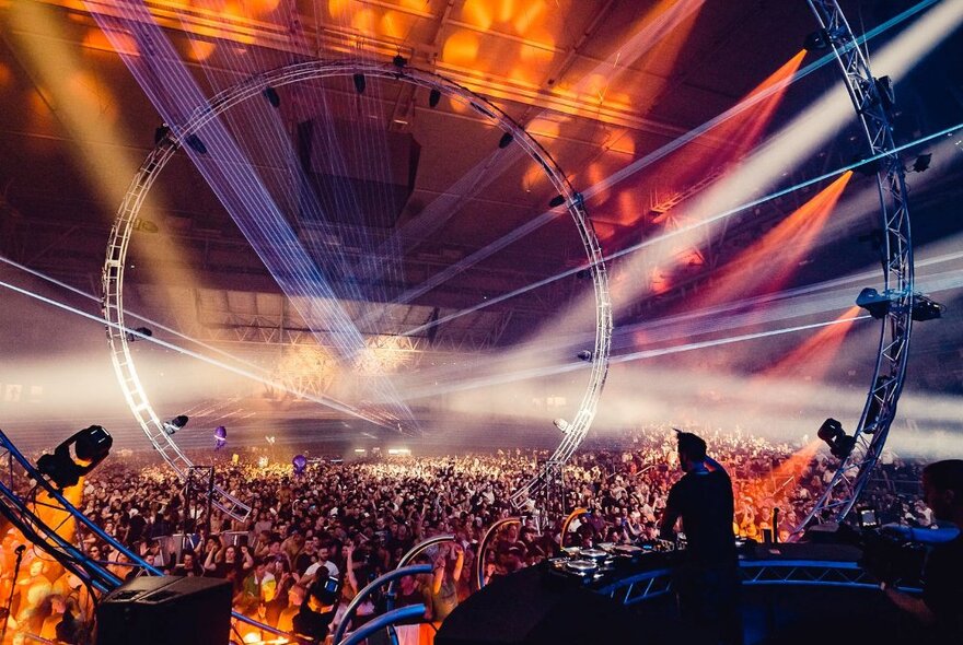 A scene from a trance music festival with a huge crowd and circular lights and projections above them. 