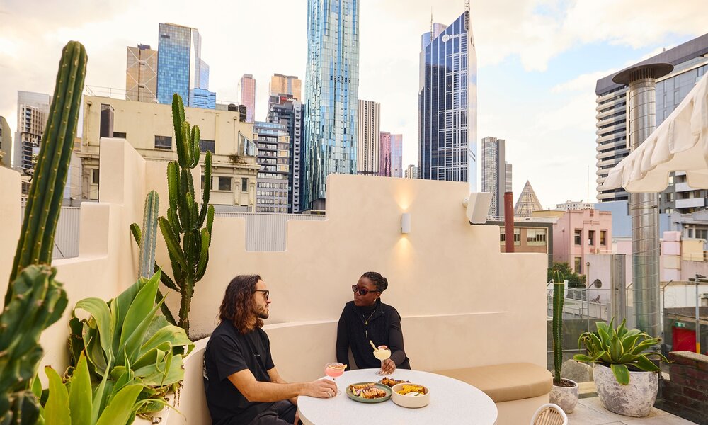 A couple dining on share plates and margaritas on a city rooftop bar with cacti.