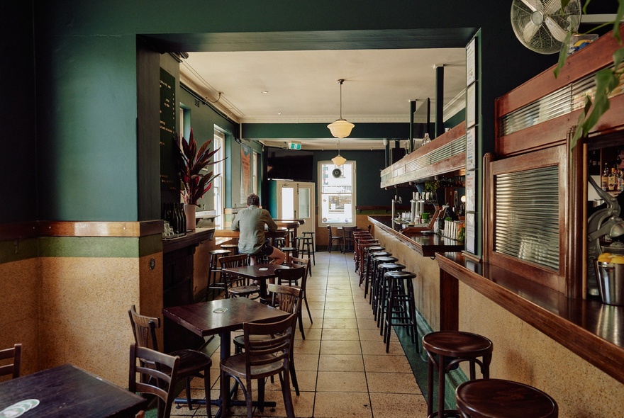 A long view down a pub bar area with stools at the bar and tables near the windows.