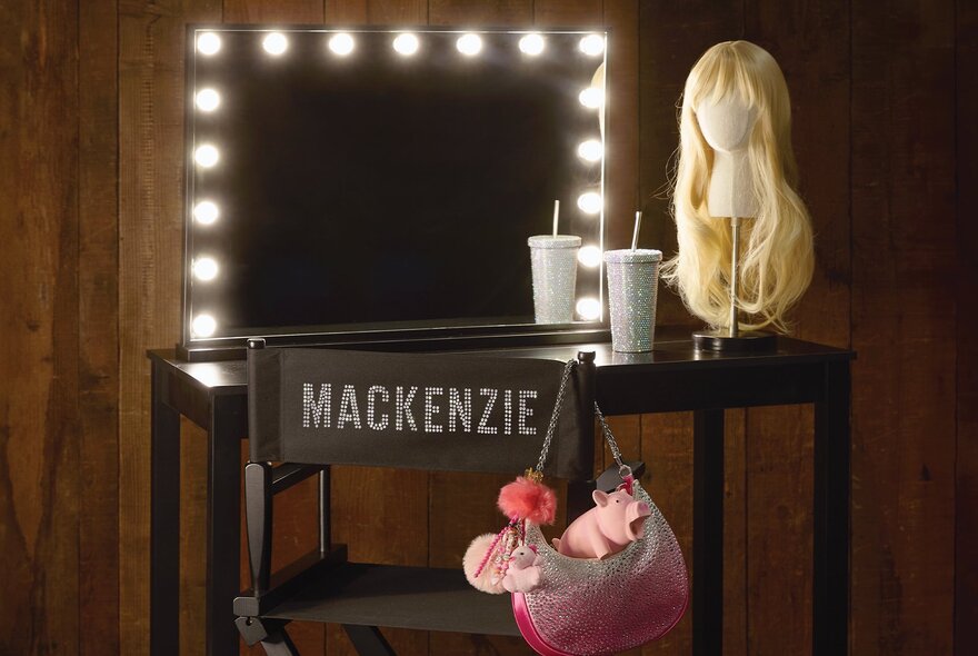 A director's chair in front of a small desk and mirror with makeup lights and a wig on a mannequin stand.