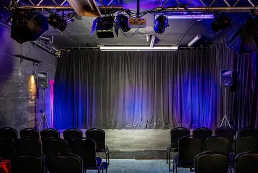 Small empty venue with rows of seating and purple-lit stage with grey curtains.