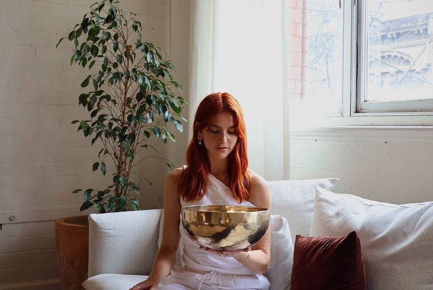 Woman seated on a couch holding a silver bowl next to a potted plant.