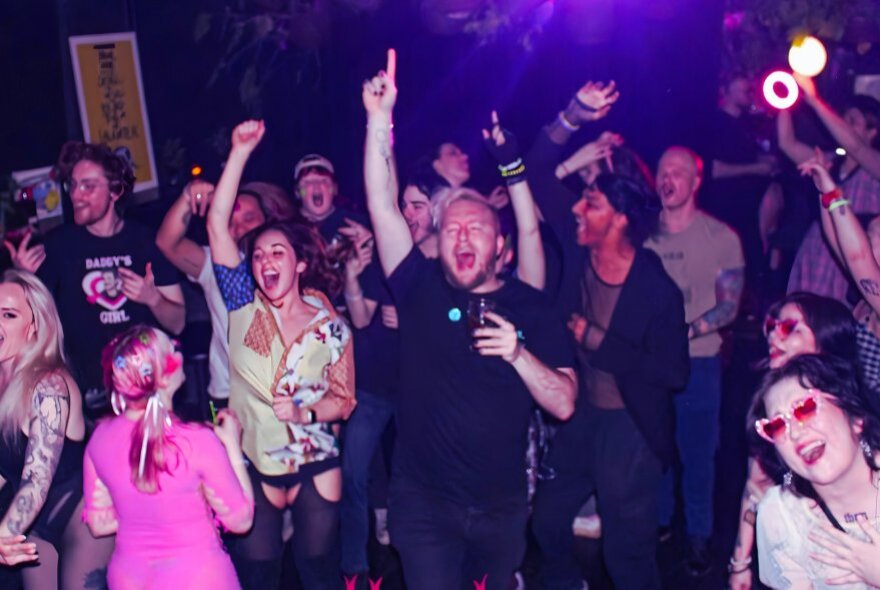 A crowd of people on a nightclub floor laughing, dancing and singing en masse, karaoke style.