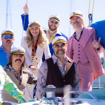 Members of Yacht Rock Revival posing on a yacht in colourful 1970s-style suits, moustaches and captains hats.