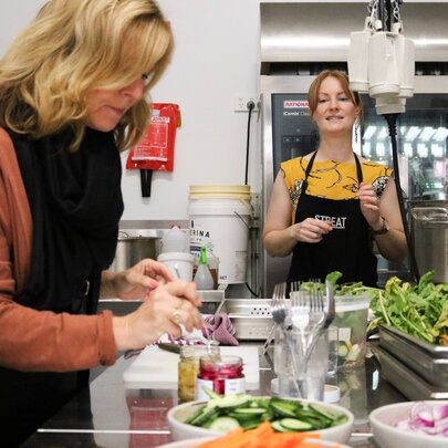 Two people in a commercial kitchen, in front of a bench filled with raw food and cooking utensils, preparing jars of stock.