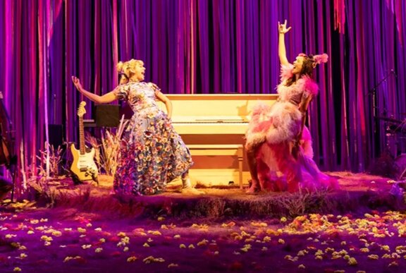 Two actors performing on a stage with purple curtains and a piano.