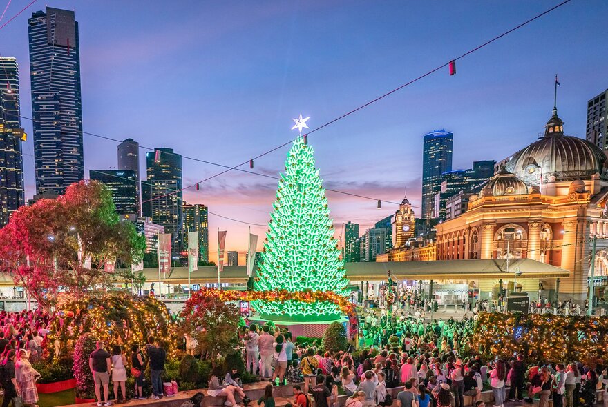a crowd of people are gathered at Melbourne's Fed Square at Christmas time with a large Christmas tree lit up in green lights